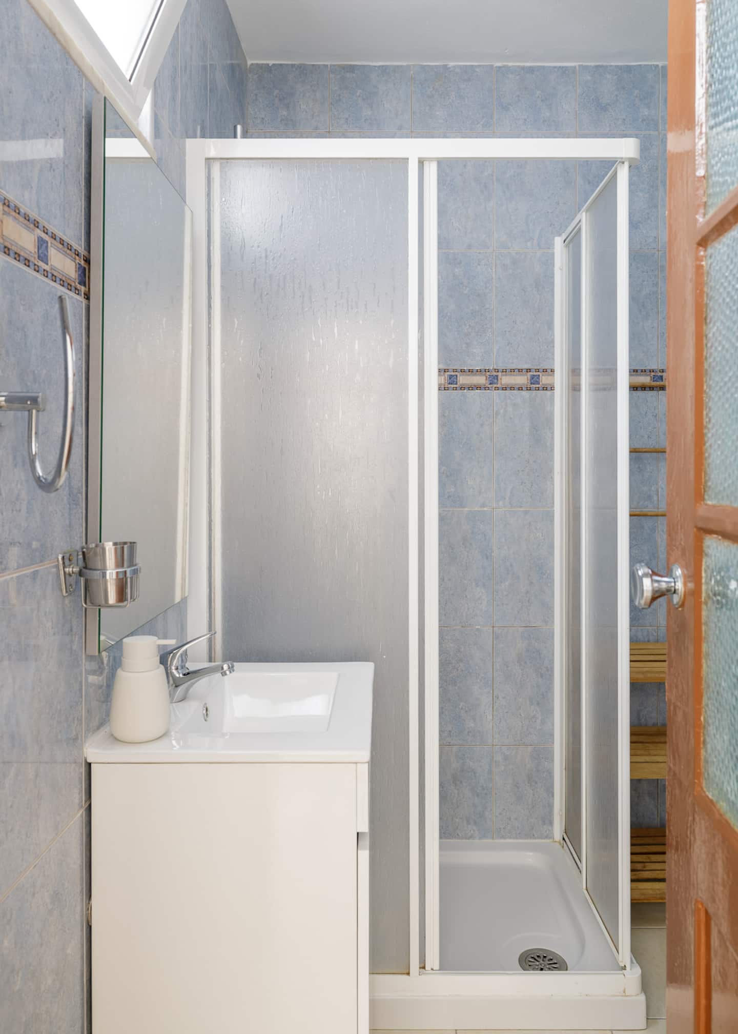 Uma casa de banho de azulejos azul-claros com uma cercadura decorada: uma cabine de duche com vidro fosco, um lavatório branco sobre um móvel branco com espelho por cima, um toalheiro de madeira em escada, e uma porta de madeira vermelha com vidros.
