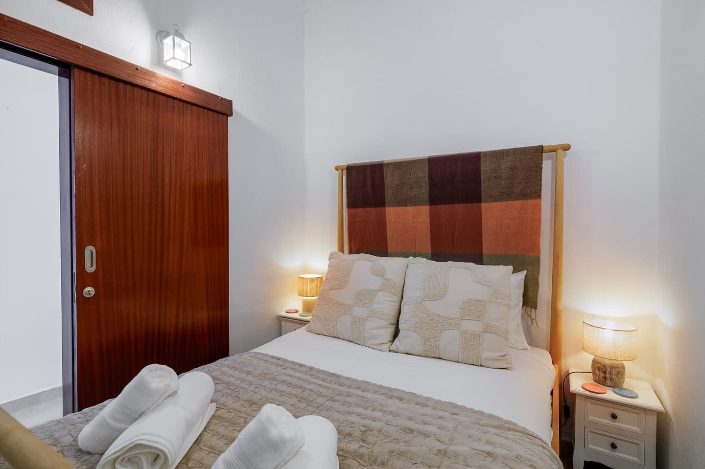 Uma cama de casal com roupa branca e uma manta bege, com toalhas dobradas em cima, uma cabeceira coberta por uma manta às riscas em tons de ferrugem, castanho e verde, candeeiros de cabeceira acesos de ambos os lados, e à esquerda uma porta de correr em madeira vermelho-escura.