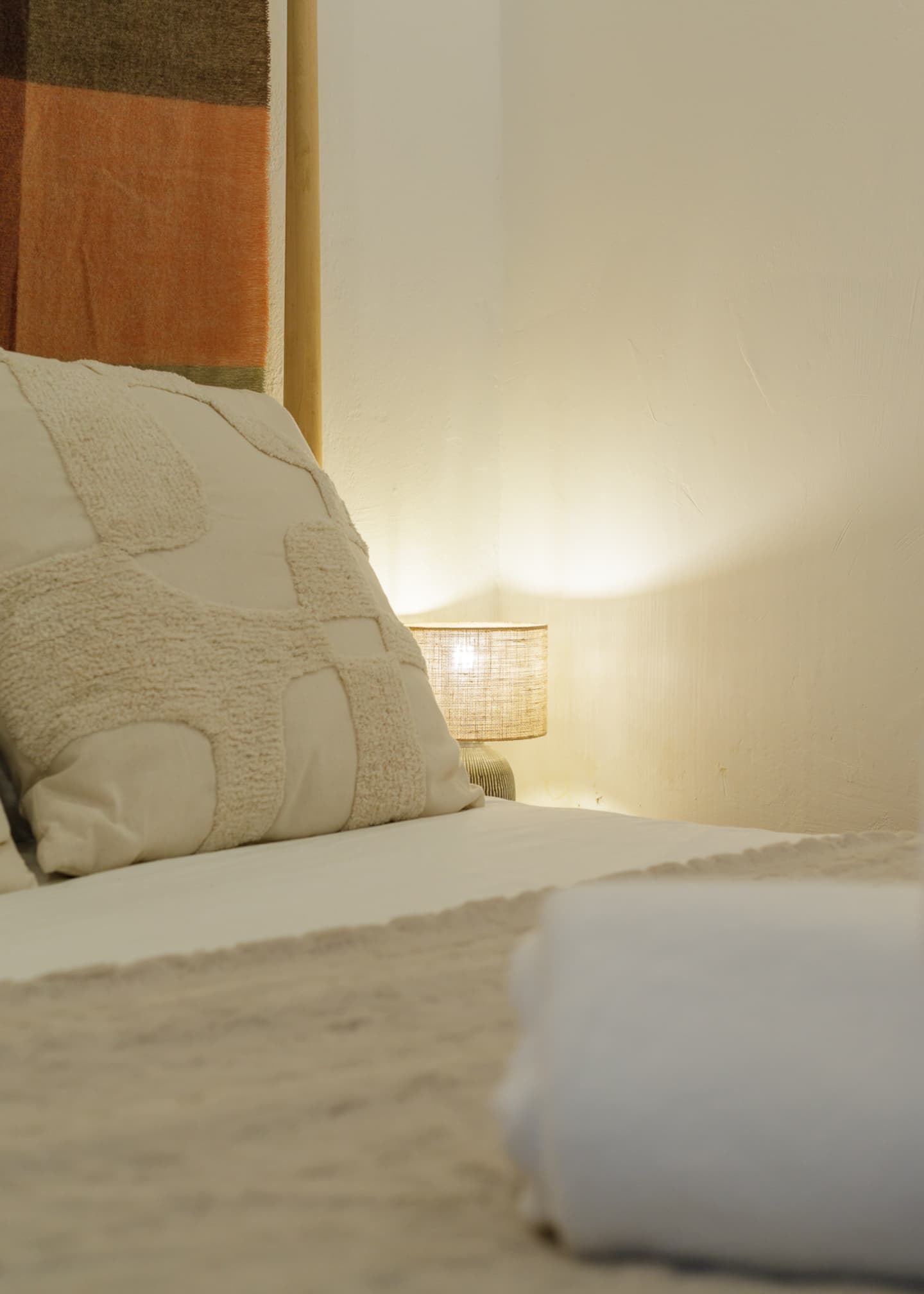 Uma almofada creme com padrão em relevo encostada à manta da cabeceira, um pequeno candeeiro de cabeceira com luz quente sobre a parede branca rebocada, e uma toalha branca dobrada em cima da cama.