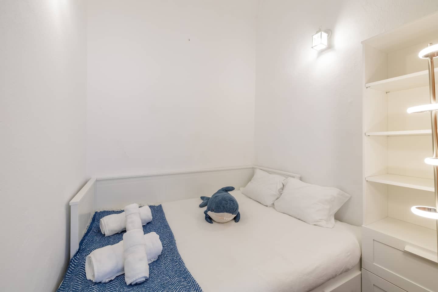 Um pequeno quarto branco: uma cama com almofadas brancas, uma manta com padrão azul e branco, toalhas brancas enroladas e um tubarão azul de peluche, um candeeiro de parede em forma de lanterna por cima, e prateleiras brancas à direita.