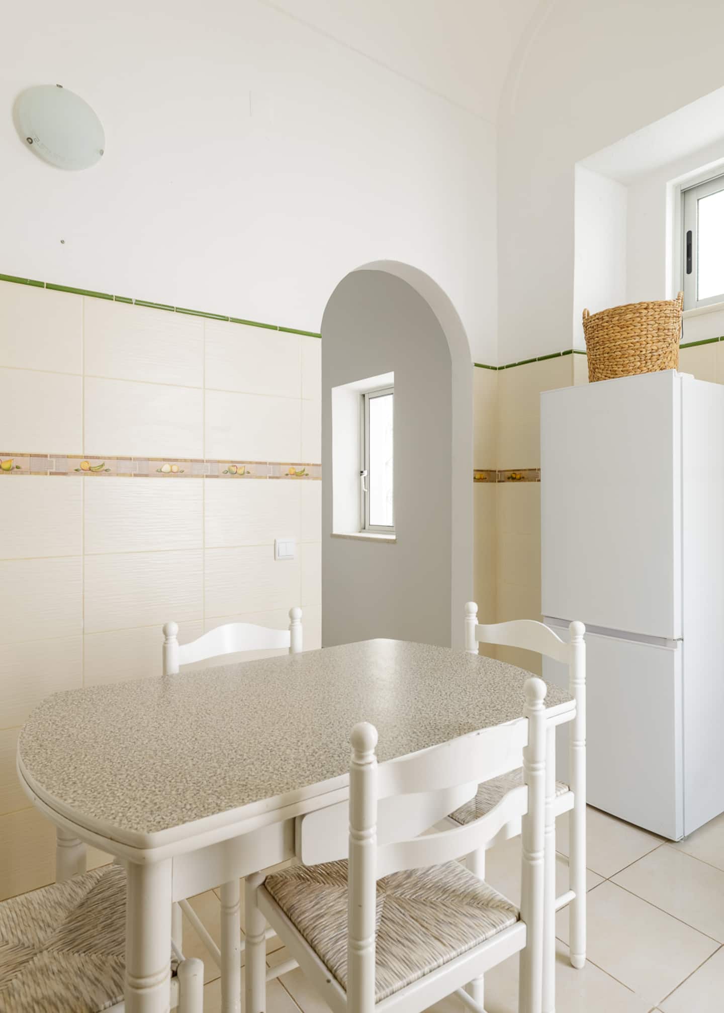Uma mesa de refeições branca com tampo mosqueado e cadeiras brancas com assento de palhinha, uma abertura em arco na parede branca com uma pequena janela ao fundo, um frigorífico combinado branco e alto com um cesto de vime em cima, e azulejos creme com um friso verde.