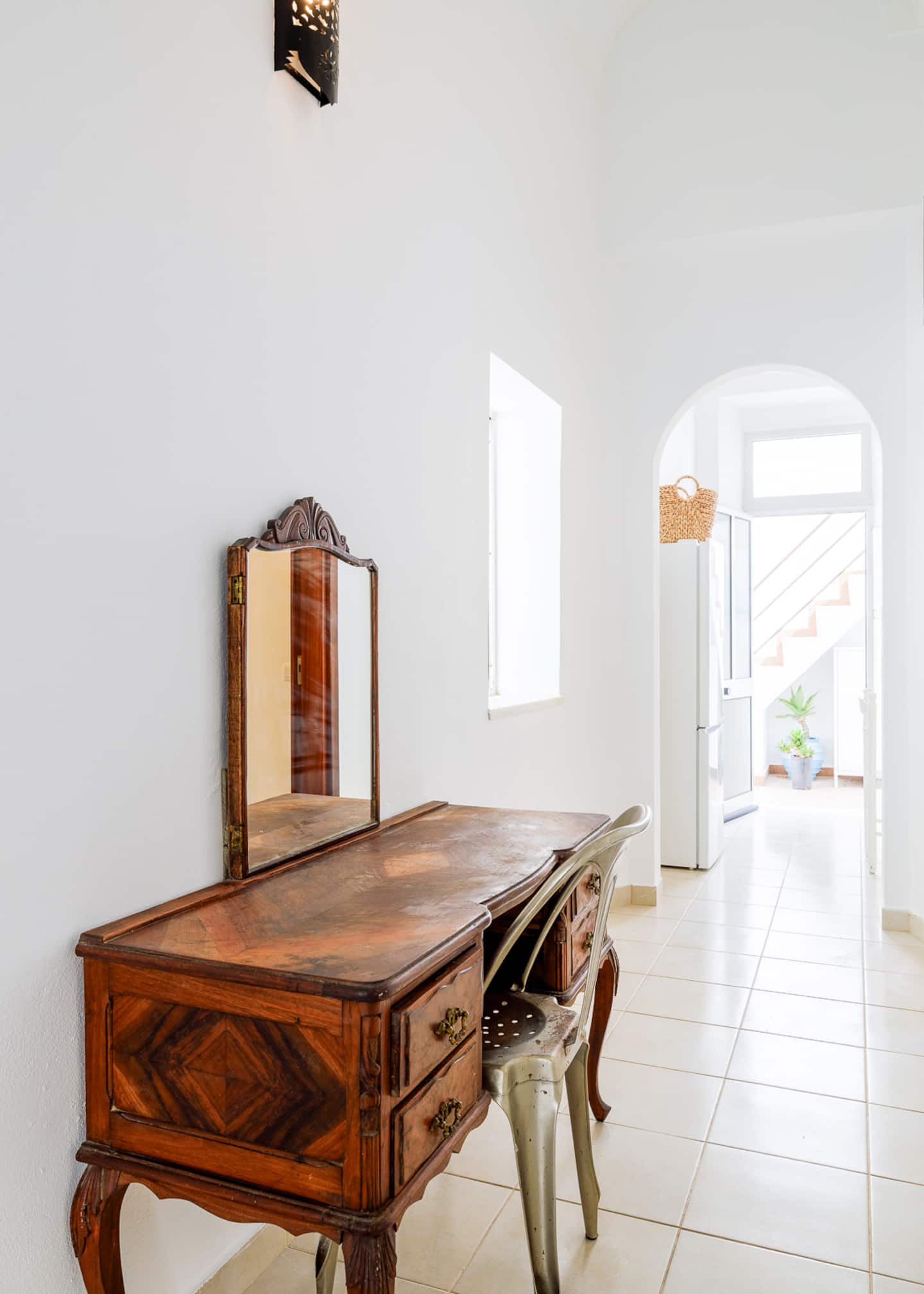 Um toucador antigo de madeira com espelho articulado e uma cadeira de café em metal, encostados a uma parede branca; para lá de um arco, o frigorífico da cozinha com um cesto de vime em cima e, através de uma porta envidraçada, a escada exterior.