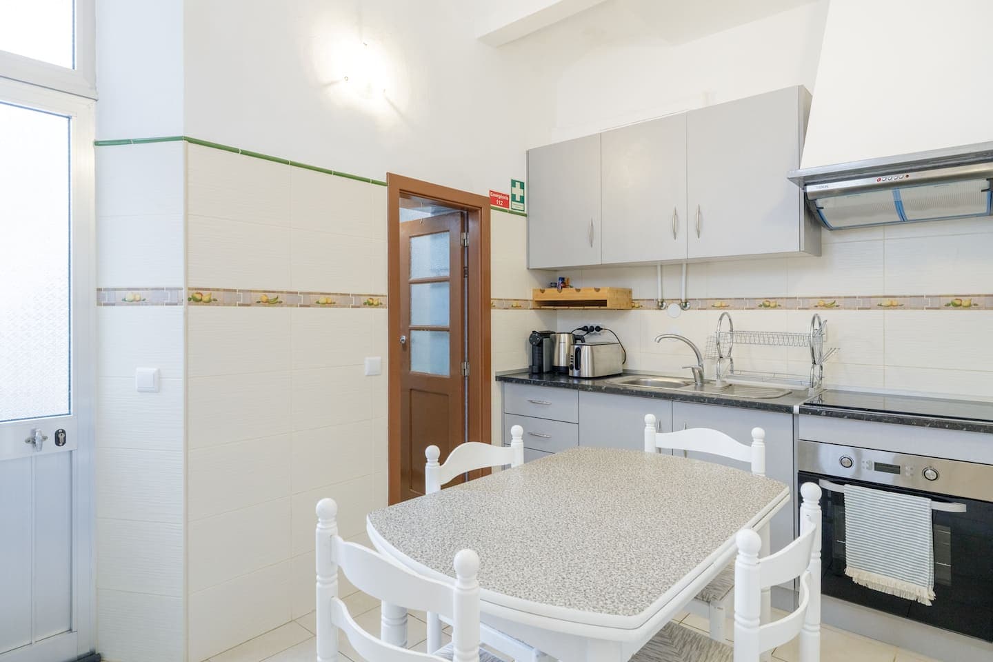 Uma cozinha com paredes de azulejo creme e uma cercadura de azulejos com frutas: armários cinzentos, lava-loiça e placa sob um exaustor em inox, um forno encastrado, uma mesa branca com quatro cadeiras brancas, e uma porta envidraçada em madeira vermelho-escura.