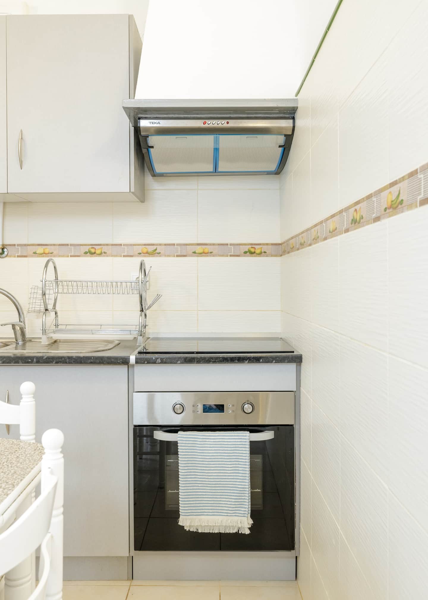 Uma placa vitrocerâmica e um forno em inox encastrados em armários cinzentos, sob um exaustor; azulejos creme com uma cercadura de limões e frutas, um escorredor de loiça junto ao lava-loiça, e um pano de cozinha às riscas no puxador do forno.