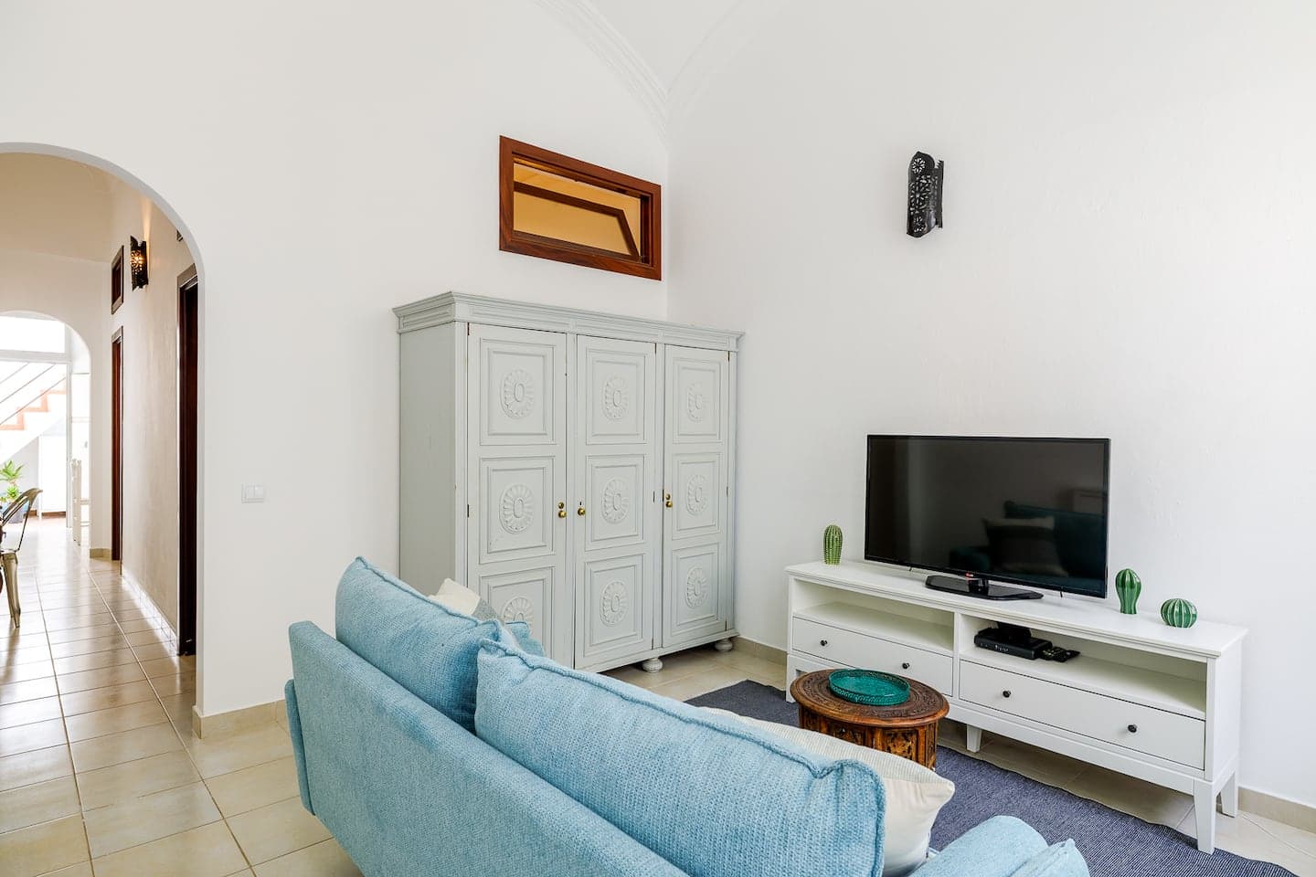 Uma sala de estar de paredes brancas e chão de mosaico bege: um sofá azul-celeste sobre um tapete cinzento, virado para uma televisão num móvel branco; atrás, um roupeiro cinzento-claro com painéis entalhados, e à esquerda uma porta em arco que dá para o corredor.