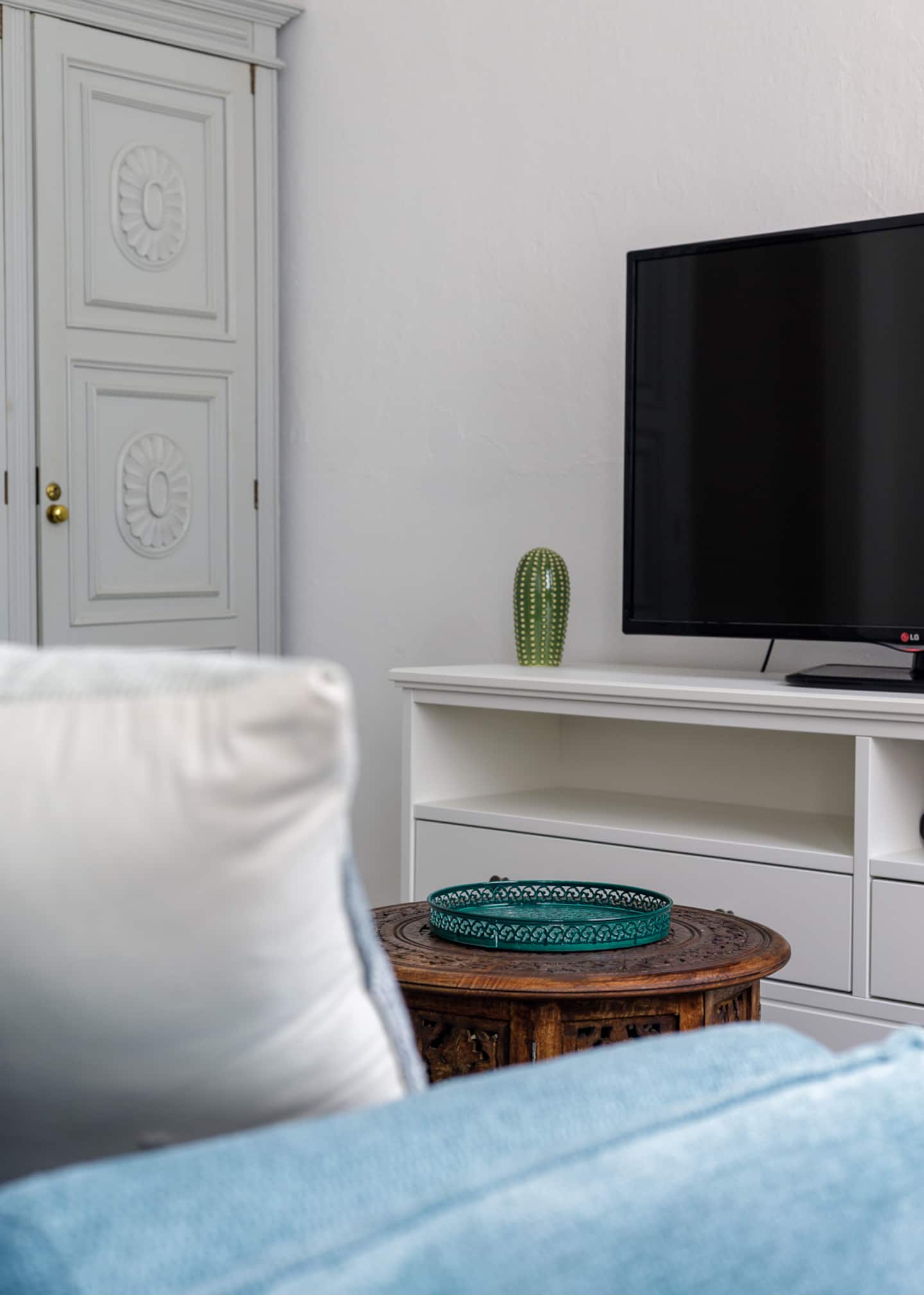 Para lá de uma almofada branca no sofá azul: uma mesa de apoio redonda em madeira entalhada, com um tabuleiro verde-azulado, a porta de um roupeiro cinzento-claro com rosetas entalhadas, um cato decorativo e uma televisão num móvel branco.