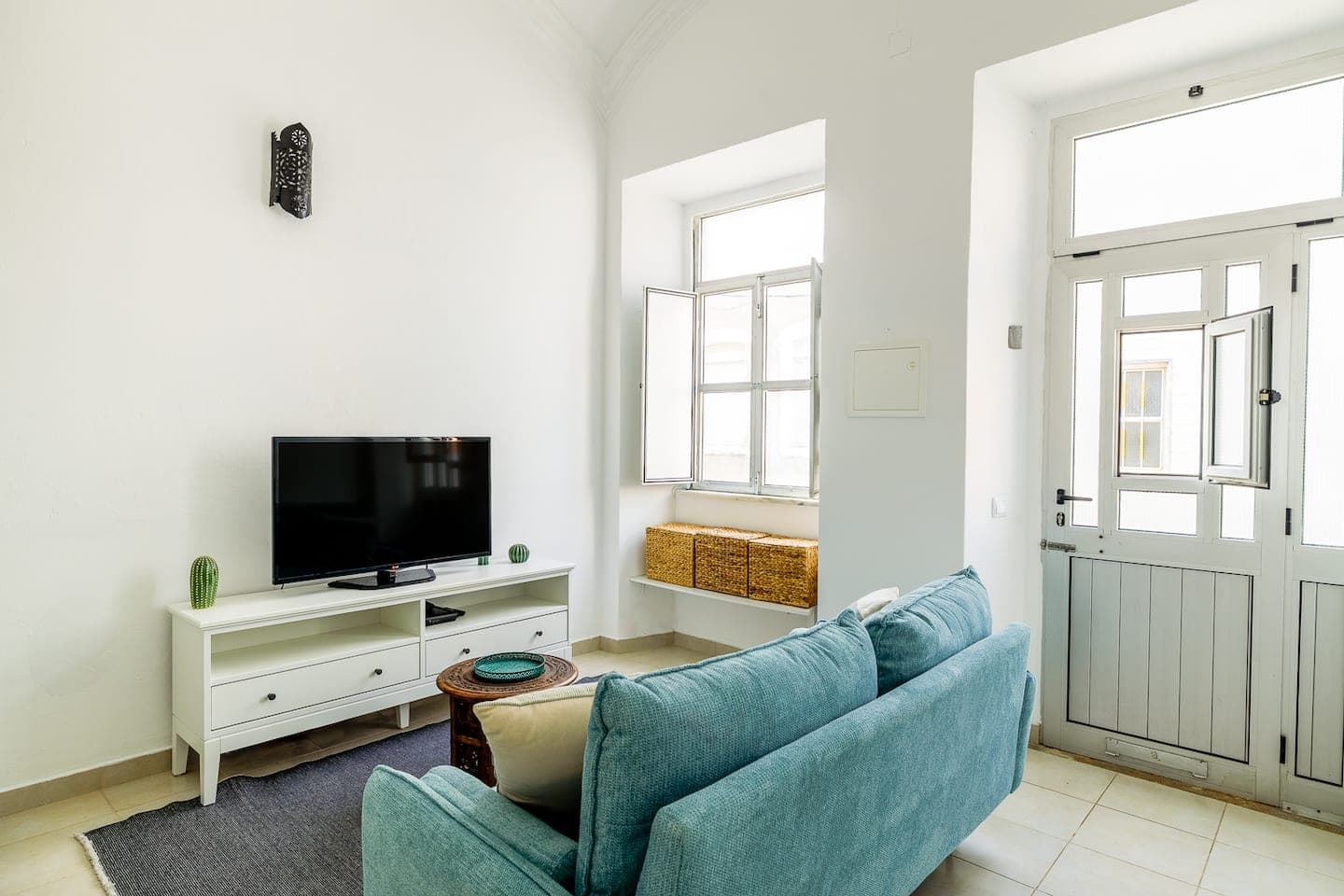 A sala de estar vista em direção à rua: uma janela funda com as portadas abertas e cestos de vime no parapeito, uma porta de entrada branca meio envidraçada, o sofá azul e uma televisão num móvel branco.