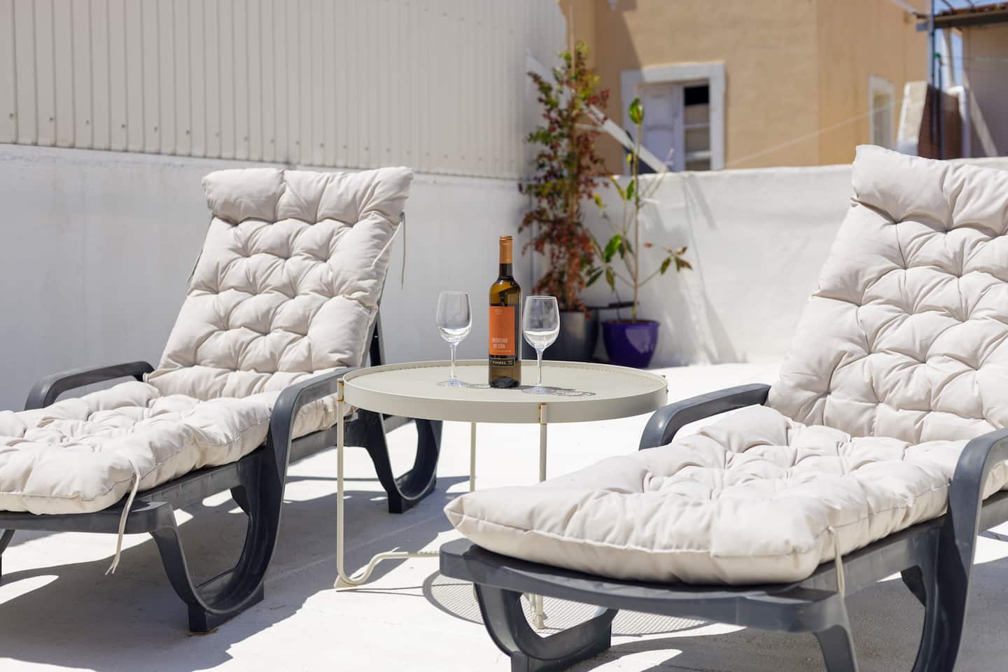 Duas espreguiçadeiras almofadadas com uma pequena mesa redonda entre elas, onde estão uma garrafa de vinho branco e dois copos, sobre o chão branco do terraço, com um arbusto num vaso, um vaso roxo e a casa amarela vizinha ao fundo.