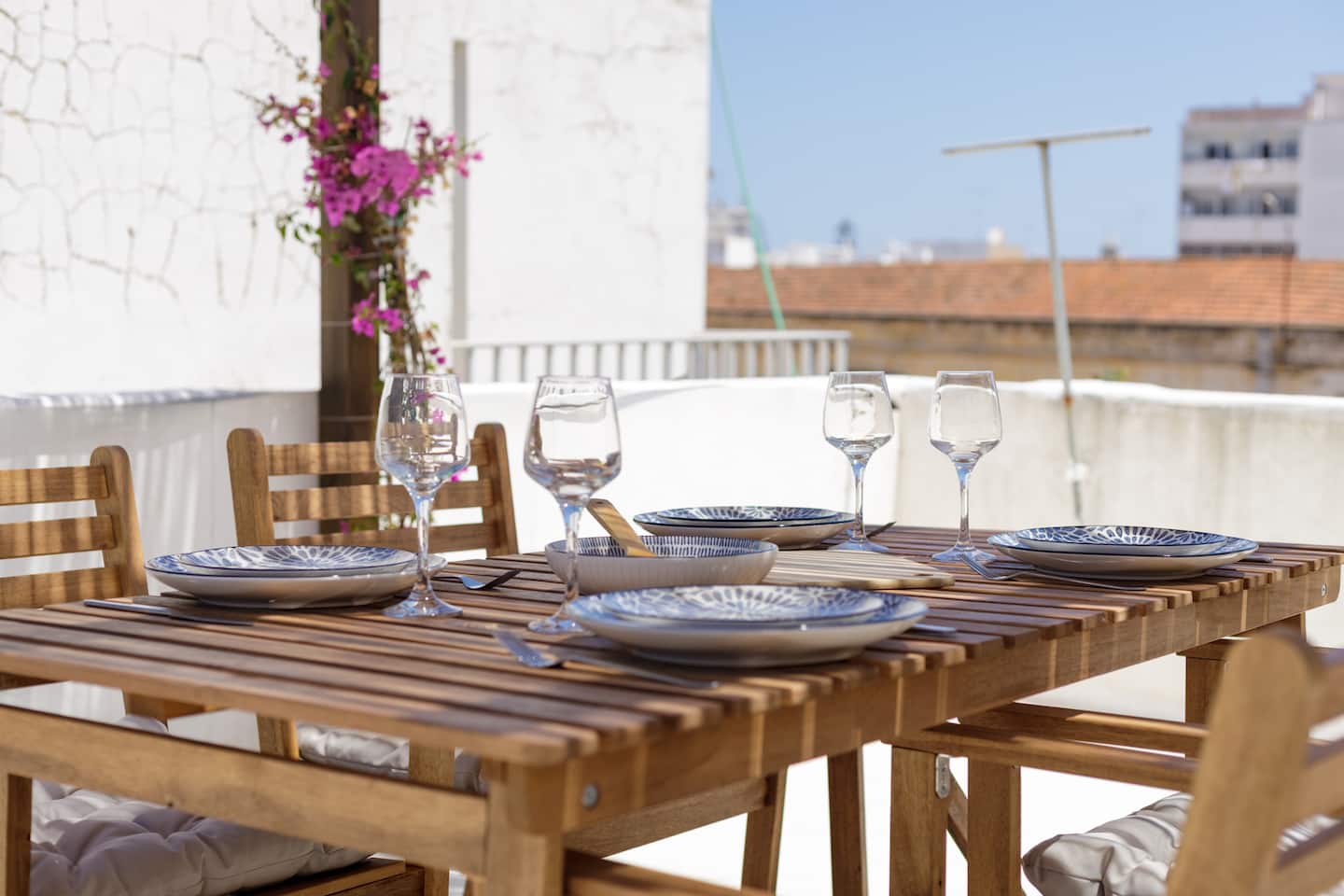 Uma mesa de ripas de madeira posta com pratos de padrão azul, taças e quatro copos de vinho, buganvília magenta sobre a parede branca por trás, e ao fundo os telhados de telha vermelha de Olhão.