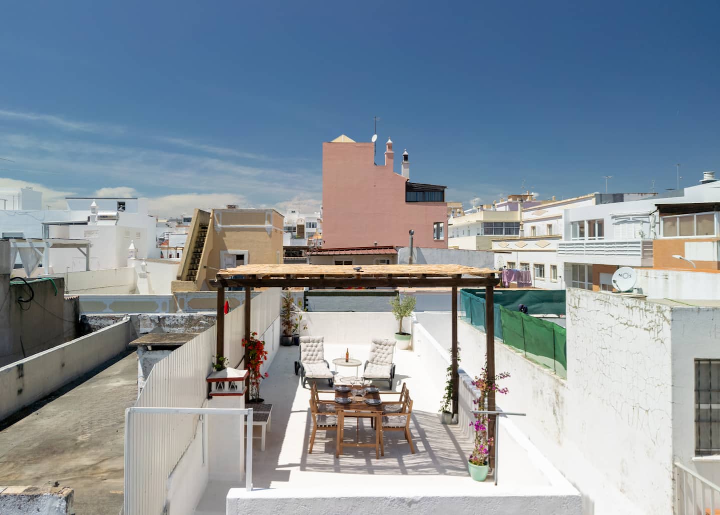 A açoteia vista de cima: a pérgula de madeira sobre a mesa de refeições, duas espreguiçadeiras e uma mesa redonda no chão branco, muros à volta, e os edifícios cor-de-rosa e brancos de Olhão sob um céu azul.
