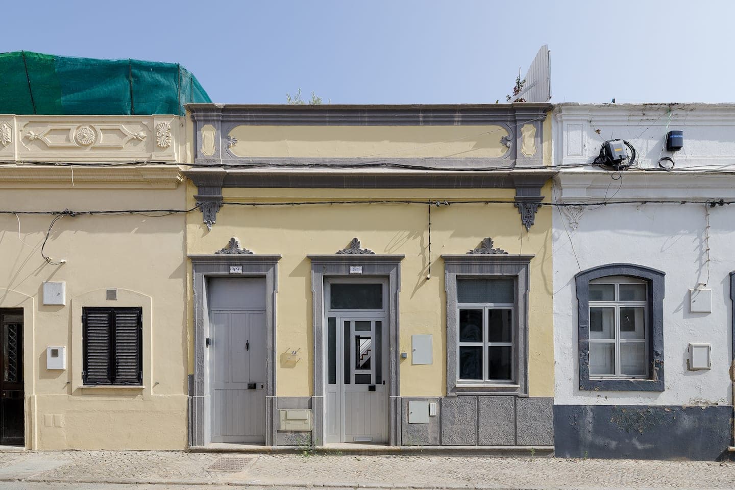 A fachada de uma casa pintada de amarelo-claro, com molduras de pedra cinzenta: duas portas, uma delas meio envidraçada, e uma janela, com uma platibanda decorada por cima, numa rua calcetada entre uma casa branca e uma cor-de-rosa.